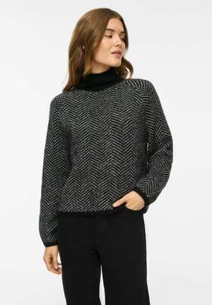Femme portant un pull à motif chevron noir et gris avec un col roulé noir et un pantalon noir, regardant vers sa gauche avec une main dans sa poche.
