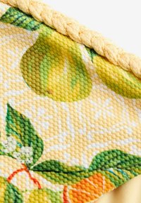 Nærbillede af tekstureret stof med gul fletkant og trykte grønne citroner, blade og appelsinfrugt på en lys baggrund.