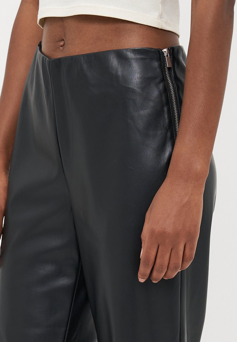 Černé kožené kalhoty s hladkou texturou, bočním zipem, přizpůsobeným střihem a minimalistickým designem. Viditelná ruka spočívající na pase.