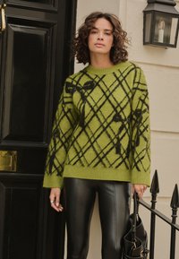Green knitted sweater with black geometric pattern, ribbed cuffs and hem, paired with black leather leggings and holding a handbag.
