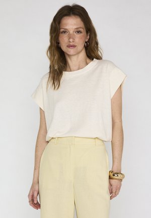 Donna con capelli castani ondulati che indossa una maglietta cremisi a maniche corte, pantaloni a vita alta giallo chiaro, orecchini a cerchio dorati, braccialetti e anelli su sfondo semplice.