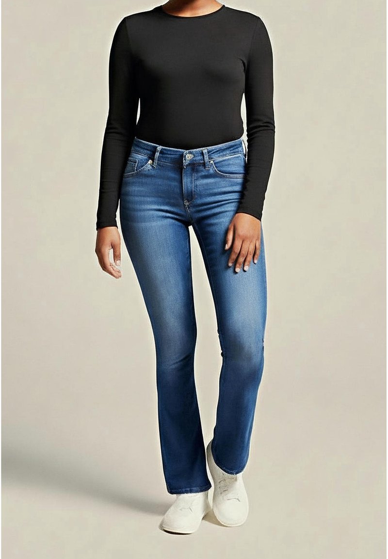 Frau in eng anliegendem, schwarzen Langarmoberteil, blauen Bootcut-Jeans und weißen Sneakers, die vor einem schlichten beigen Hintergrund steht.