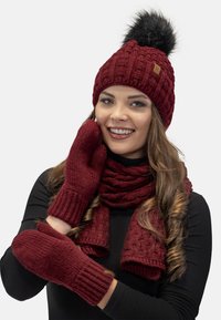 Cappello a maglia bordeaux con un pom-pom in pelliccia sintetica nera, guanti coordinati e una sciarpa texturizzata. Il tessuto presenta un motivo a trecce.