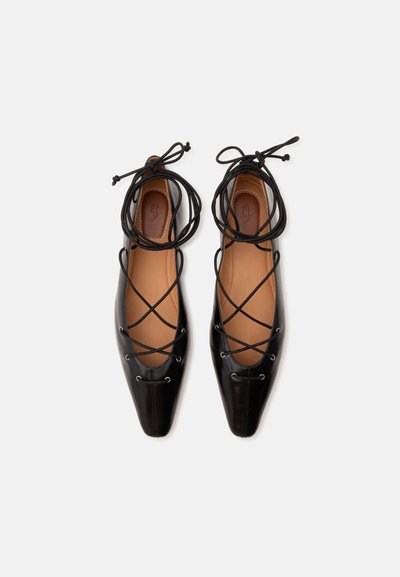Paire de ballerines en cuir noir avec bouts pointus et brides à lacets croisées sur fond blanc.