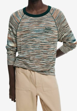 Person trägt einen mehrfarbigen, gestreiften Pullover mit langen Ärmeln und dunkelgrünem Kragen sowie beige Hose, steht mit einer Hand hinter dem Rücken.