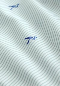Tela verde clara con sutiles rayas onduladas, que presenta bordados de flamencos azules como detalle destacado. Textura suave y apariencia ligera.