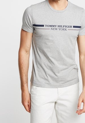 Hombre con una camiseta ajustada gris claro Tommy Hilfiger New York y pantalones blancos, mostrado desde el cuello hasta las caderas sobre un fondo liso.