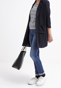 Svart lång cardigan med en ficka, randig skjorta, blå jeans och vita sneakers. Tillbehören inkluderar en svart tote-väska.