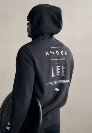 Haut à capuche noir avec une poche kangourou à l'avant et un grand logo au dos. Tissu doux, présentant une liste de villes et d'événements en texte blanc et vert.