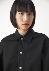 Veste noire à boutons, avec un col, dotée de boutons-pression en argent et d'un tissu texturé. Gros plan sur le design avant.