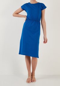 Kvinna i en knälång, kunglablå klänning med korta ärmar, med ena handen på höften, iförd sandaler med genomskinliga remmar, stående mot en enfärgad bakgrund.