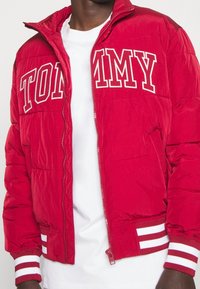 Röd vadderad jacka med vit "TOMMY"-text, dragkedjestängning, sidofickor och randiga manschetter. Släta ytor med quiltade detaljer.