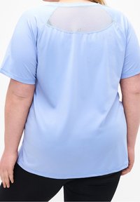 Mujer vestida con una camiseta deportiva de manga corta azul claro, con panel de malla en la parte superior de la espalda, combinada con pantalones negros, de espaldas.