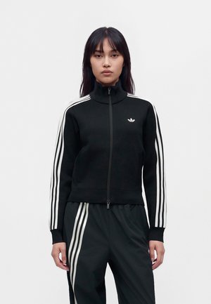 Жена представя черен спортен екип Adidas с бели ивици и яке с цип на изчистен бял фон.