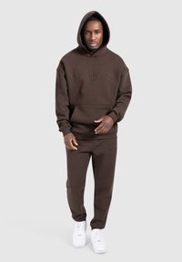 Brauner Hoodie mit einer Fronttasche und eingeprägtem Logo, kombiniert mit passenden Sweatpants. Beide aus einem weichen, strukturierten Stoff. Weiße Sneakers.