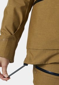 Chaqueta Helly Tech marrón con un patrón texturizado, cordón ajustable en el dobladillo y detalle de bolsillo con cremallera en negro.