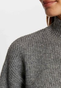 Pull en gris côtelé avec un col montant, présentant une texture douce et un léger brillant, mettant en valeur le motif tricoté et les détails du col.