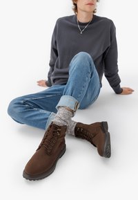 Pruunid sametist pahkluu saapad lace-up kinnitusega, kummist tallaga ja tekstureeritud detailidega, koos siniste teksade ja mustriliste kudum socksidega.