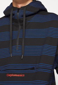 Felpa nera con strisce orizzontali blu, zip frontale e logo rosso "Cleptomanicx". Presenta coulisse e tessuto morbido.