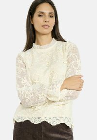 Blusa in pizzo di colore avorio con orlo a onde, ricami floreali e scollatura arricciata. Maniche lunghe in tessuto trasparente con dettagli in pizzo intricato.