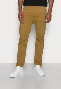 Cargo-Hosen in Khaki-Farbe, mit mehreren seitlichen Taschen, einer schmalen Passform und einem atmungsaktiven Baumwoll-ähnlichen Material. Getragen mit weißen Sneakers.