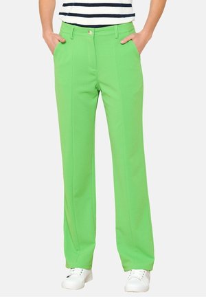 Osoba ubrana w jaskrawozielone spodnie o prostych nogawkach, białe trampki i biały top z czarnymi poziomymi paskami, z rękami w kieszeniach.