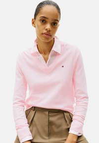 Helles rosa V-Ausschnitt-Pullover mit gerippten Bündchen, über ein weißes Hemd mit Kragen getragen. Besitzt ein kleines Logo auf der Brust und eine glatte Textur.