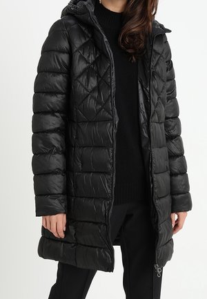 Femme portant un manteau matelassé noir rembourré, longueur genoux, avec une capuche, ouvert sur un pull noir et un pantalon noir.