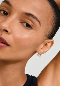 Boucles d'oreilles en or en forme de divers styles : une barre droite, un design tordu et un petit accent en perles. La personne qui les porte a les cheveux coiffés en arrière.