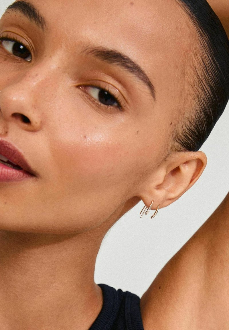 Boucles d'oreilles en or en forme de divers styles : une barre droite, un design tordu et un petit accent en perles. La personne qui les porte a les cheveux coiffés en arrière.