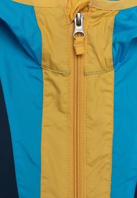 Gul och turkos jacka med en central dragkedja som har ett silver YKK-drag. Slät nylontextur med kontrasterande sömmar synliga.