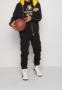 Svart träningsdräkt med gula accenter och Los Angeles Lakers-logotyp; svarta cargo-byxor; vita och röda Puma-sneakers; håller en basketboll.