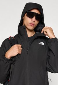 Μαύρο ελαφρύ μπουφάν με κουκούλα, φερμουάρ κλεισίματος και ελαστικά μανίκια. Διαθέτει το λογότυπο "The North Face" στο στήθος και εμφανή λουρί σακιδίου πλάτης.