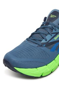 Modrá športová obuv s priedušným sieťovaným zvrškom, zelenou podrážkou a textúrovanými detailmi. Obsahuje výrazný šnurovací dizajn a akcenty loga.
