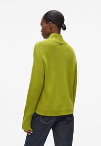 Grüner gerippter Pullover mit hohem Kragen, langen Ärmeln und einem dezenten Markenakzent auf der Rückseite. Strukturierter Strickstoff.