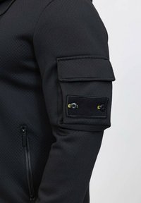 Veste à capuche noire avec un tissu texturé, poche zippée sur le côté et poche à rabat avec fermeture à bouton sur le haut du bras.