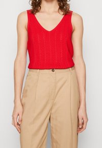 Červený pletený top bez rukávů s hlubokým výstřihem do V, s texturovaným vzorem, v kombinaci s béžovými kalhotami s vysokým pasem.