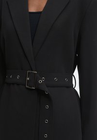 Μαύρο ραμμένο σακάκι φορεμένο με ταιριαστή ζώνη που διαθέτει μεταλλικά ρουμπάκια και ορθογώνια ασημένια πόρπη πάνω από μαύρο ριπ τοπ.