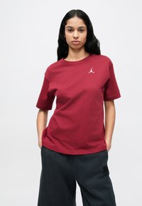 Μπεζ πουκάμισο από βαμβάκι σε μπορντό, με κοντά μανίκια, στρογγυλή λαιμόκοψη και λευκό λογότυπο Jordan στο στήθος. Συνδυάζεται με σκούρο παντελόνι.