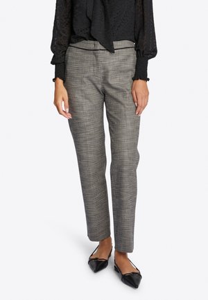 Femme portant un pantalon à carreaux gris, une blouse noire transparente à manches longues et des ballerines noires à bout pointu, debout sur fond blanc.