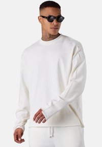 Homme portant des lunettes de soleil rondes noires et un sweat-shirt à manches longues blanc, ainsi que des pantalons blancs assortis, se tenant contre un fond uni.