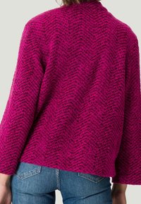 Fuchsiový strukturovaný svetr se zigzagovým vzorem, širokými rukávy a volným střihem, v kombinaci s modrými džíny. Zadní pohled ukazuje designové detaily.