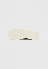 Suela de zapatilla en goma beige con un patrón texturizado, que presenta una impresión de logo y ranuras para tracción. Superficie lisa con líneas curvas.