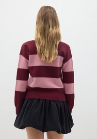 Gestreifter Pullover in Burgunderrot und Pink, aus weichem Strickmaterial. Verfügt über lange Ärmel und einen taillierten Bund, kombiniert mit einem schwarzen Rock.