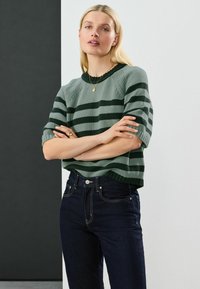 Maglione lavorato a maglia verde con strisce verde scuro, scollatura rotonda e maniche corte, abbinato a jeans in denim scuro con cuciture in risalto.