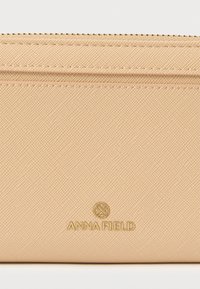 Bēšas krāsas teksturēts maks ar gludu virsmu, zelta toni apzīmēts logotips, šūtas detaļas un plāns dizains ērtai pārnēsāšanai.