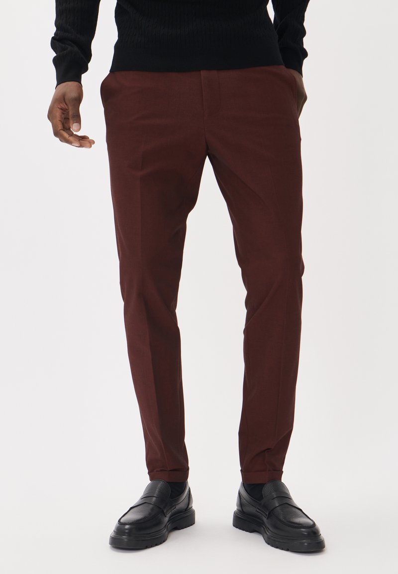 Pantalones burgundy a medida, hechos de una tela suave, con un diseño de corte ajustado y bolsillos frontales. Combinados con zapatos de cuero negros.