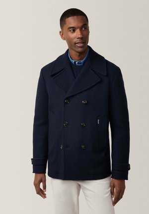 Abrigo de lana azul marino de doble botonadura con grandes solapas, botones decorativos y un bolsillo lateral, combinado con una camisa azul con cuello por debajo.