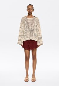 Beige gestrickter Pullover mit weiten Ärmeln und offenem Muster, über einem kastanienbraunen Kleid getragen. Model trägt rosa Rüschensandalen und steht vor einem weißen Hintergrund.