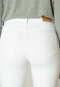 Witte denim jeans met achterzakken. Materiaal lijkt soepel en elastisch. Een tan leren label is bevestigd aan de tailleband. Boven een grijze shirt.